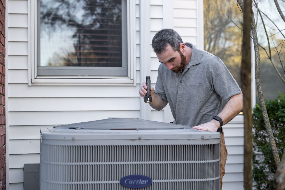 Technician inspecting an outdoor AC condenser beside a home with a flashlight